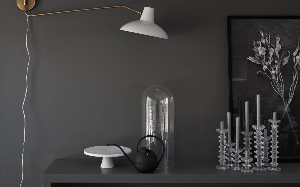 Mässingsfärgad lampa med vit hatt hängandes från grå vägg, sträcker sig över grå sideboard med smakfulla detaljer